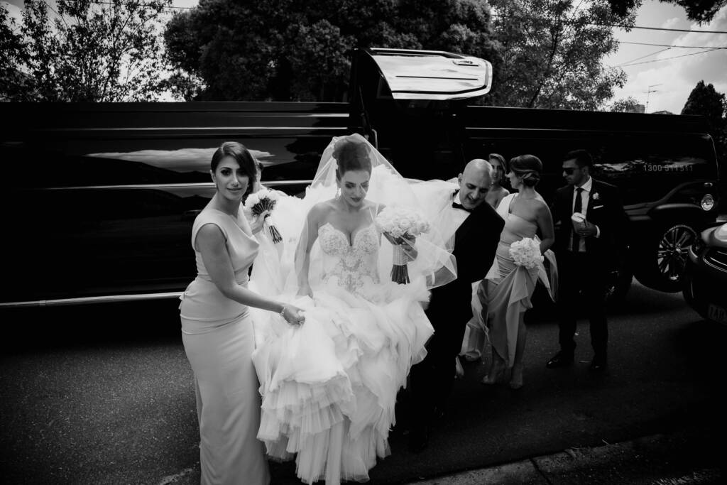 A bridesmaid gathers Danielle's ruffled skirt beside the wedding car before the ceremony