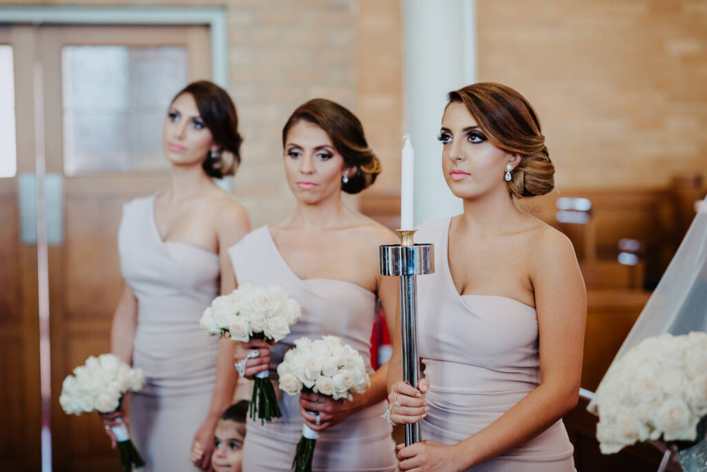 Three bridesmaids stand with bouquets during the ceremony, one holding an unlit candle