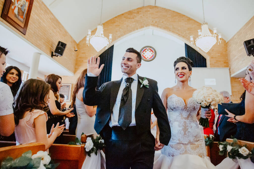 Michael waves as he and Danielle walk back down the aisle at St Aphrem Syrian Orthodox Church