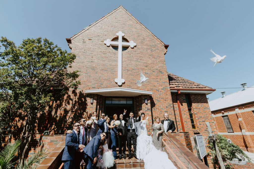 White birds fly above the wedding party outside St Aphrem Syrian Orthodox Church