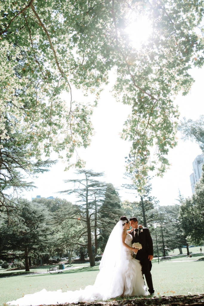 Danielle and Michael lean together beneath sunlit trees at Carlton Gardens