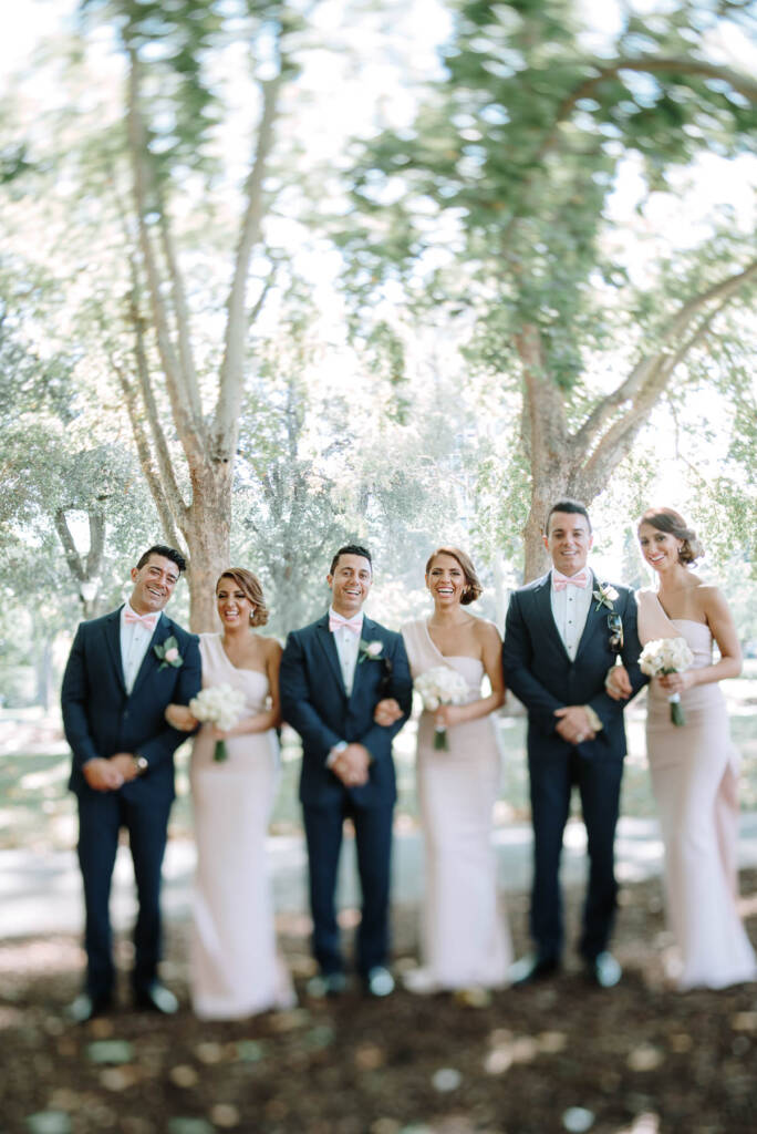 Three bridesmaids and three groomsmen pose arm in arm beneath trees at Carlton Gardens
