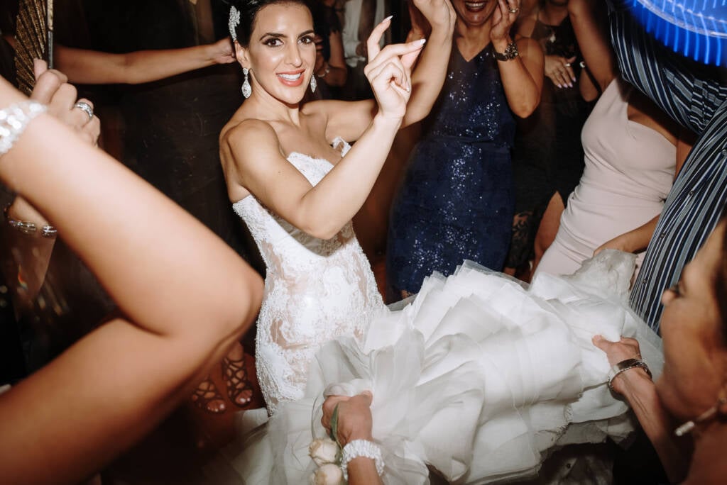 Danielle raises her hands while guests hold up the ruffled skirt of her gown