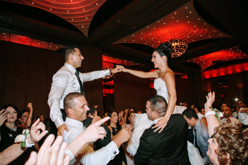 Danielle and Michael reach towards each other from guests' shoulders at Palladium