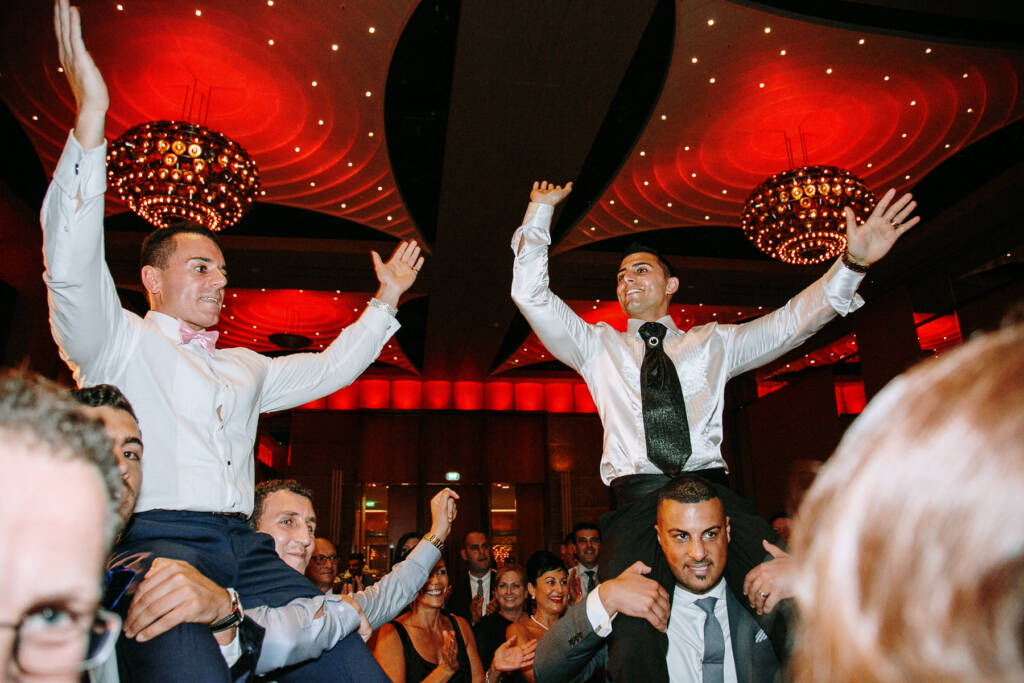 Michael and a groomsman ride guests' shoulders beneath red-lit chandeliers at Crown Palladium