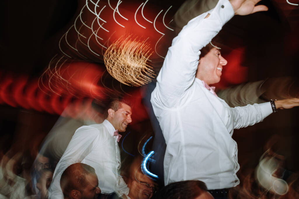 Two groomsmen ride guests' shoulders amid chandelier light trails at Palladium