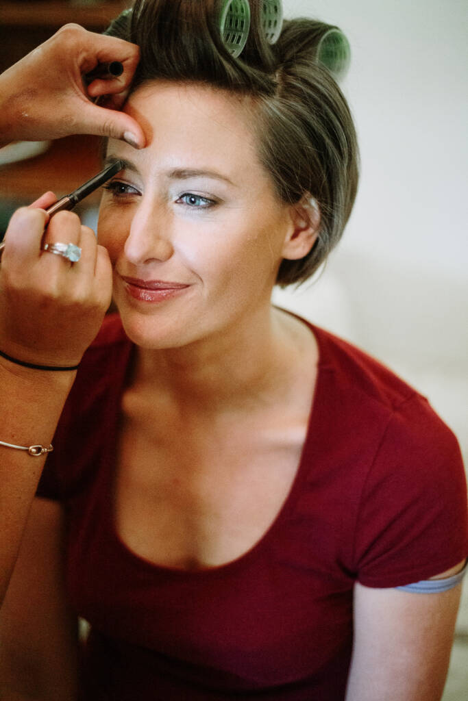 Camilla smiles in green rollers as a makeup artist shapes her brow with a small brush.