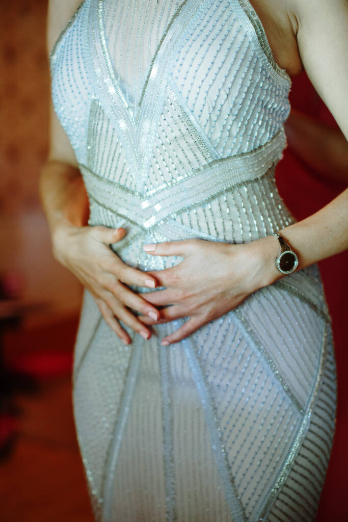 Detail of hands folded at the waist of the beaded gown, a slim silver watch on the wrist.