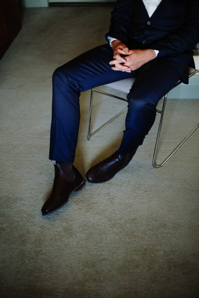 Detail of Jason's navy suit and dark boots as he sits with his hands clasped.