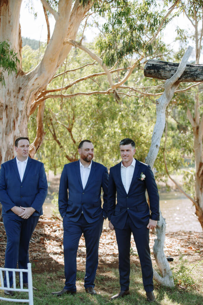 Jason and two suited companions wait beside the timber ceremony arbour and lake at Flowerdale.