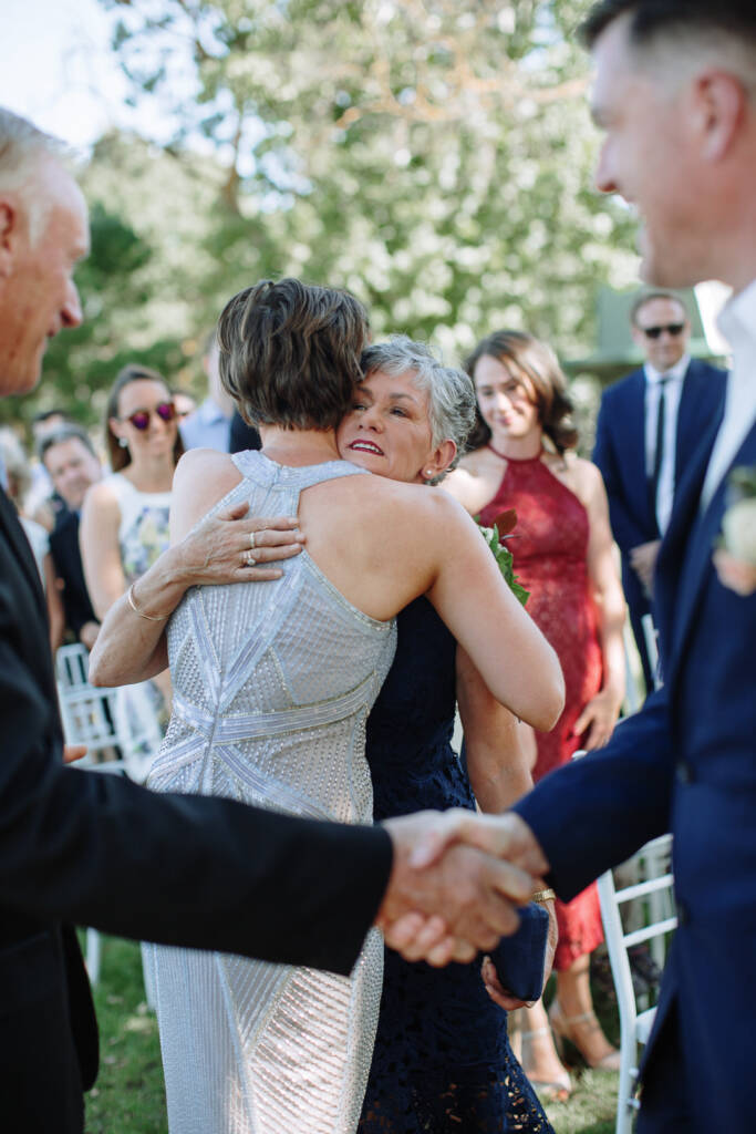 At the front of the ceremony Camilla hugs a guest in navy lace as Jason shakes hands.