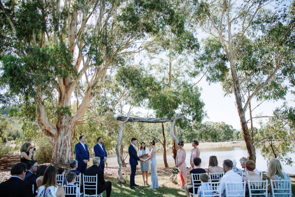 Wide view of Camilla and Jason's lakeside ceremony beneath a timber arbour at Flowerdale Estate.