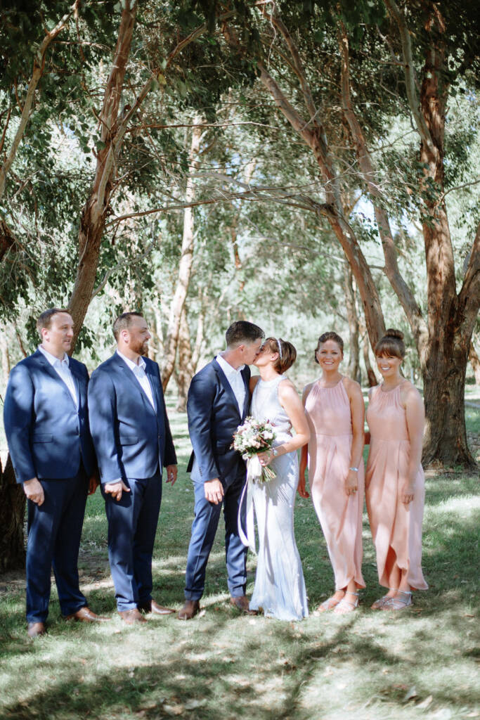 Camilla and Jason kiss in a wedding-party portrait beneath trees at Flowerdale.