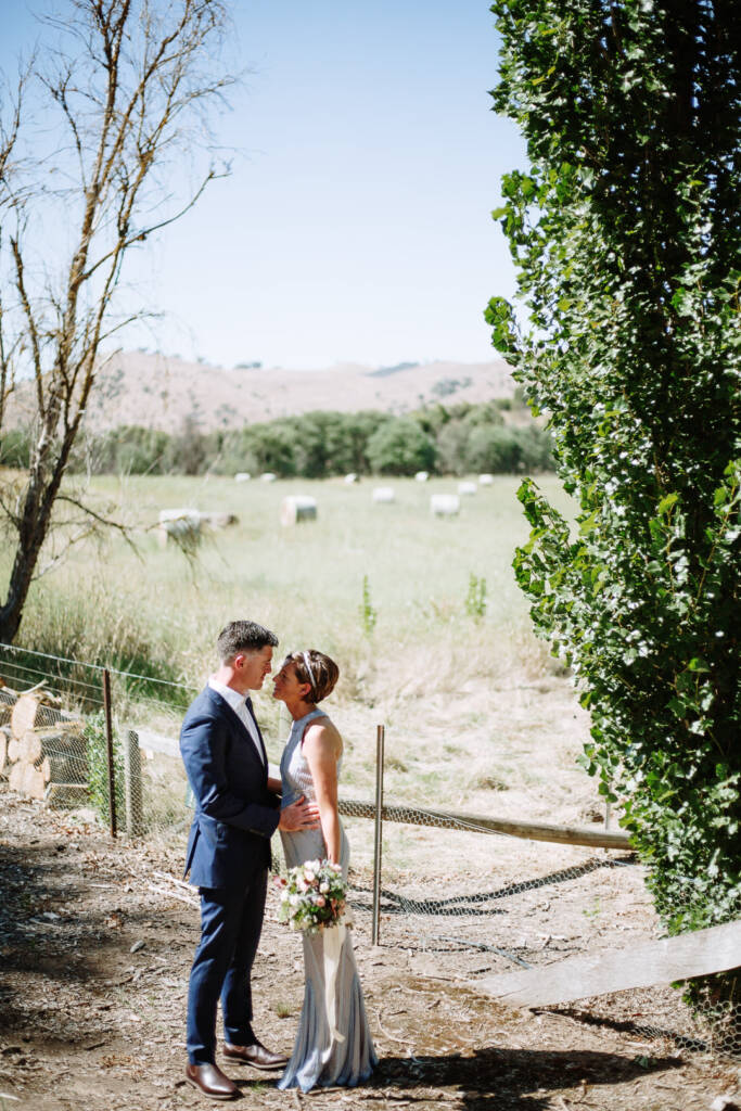 Camilla and Jason embrace beside a paddock fence with hay bales beyond at Flowerdale Estate.
