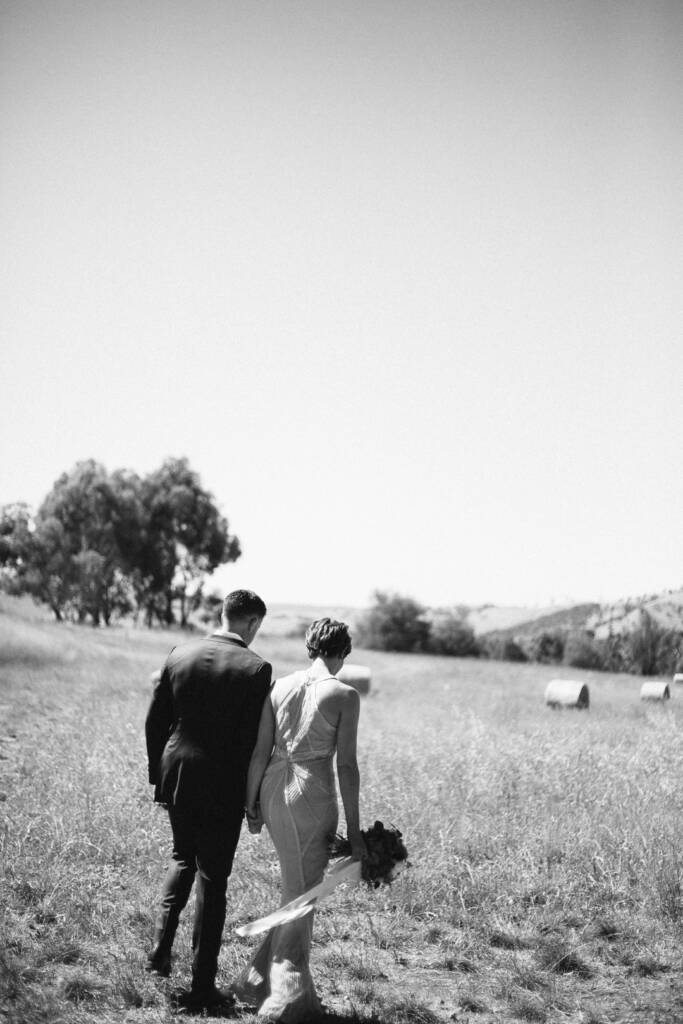 Black-and-white photo of Camilla and Jason walking hand in hand towards hay bales at Flowerdale Estate.