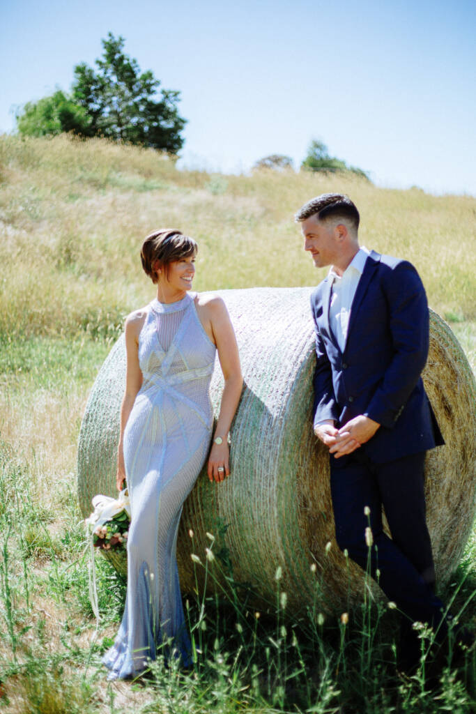 Camilla and Jason smile towards each other while leaning against a hay bale at Flowerdale Estate.