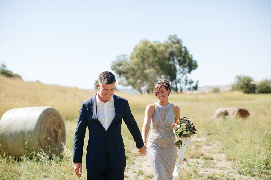 Camilla and Jason smile while walking hand in hand between hay bales at Flowerdale Estate.