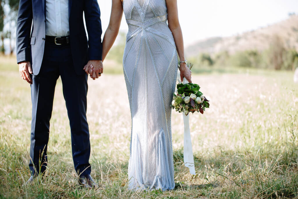 Close detail of Camilla and Jason's joined hands, beaded gown and ribboned bouquet.