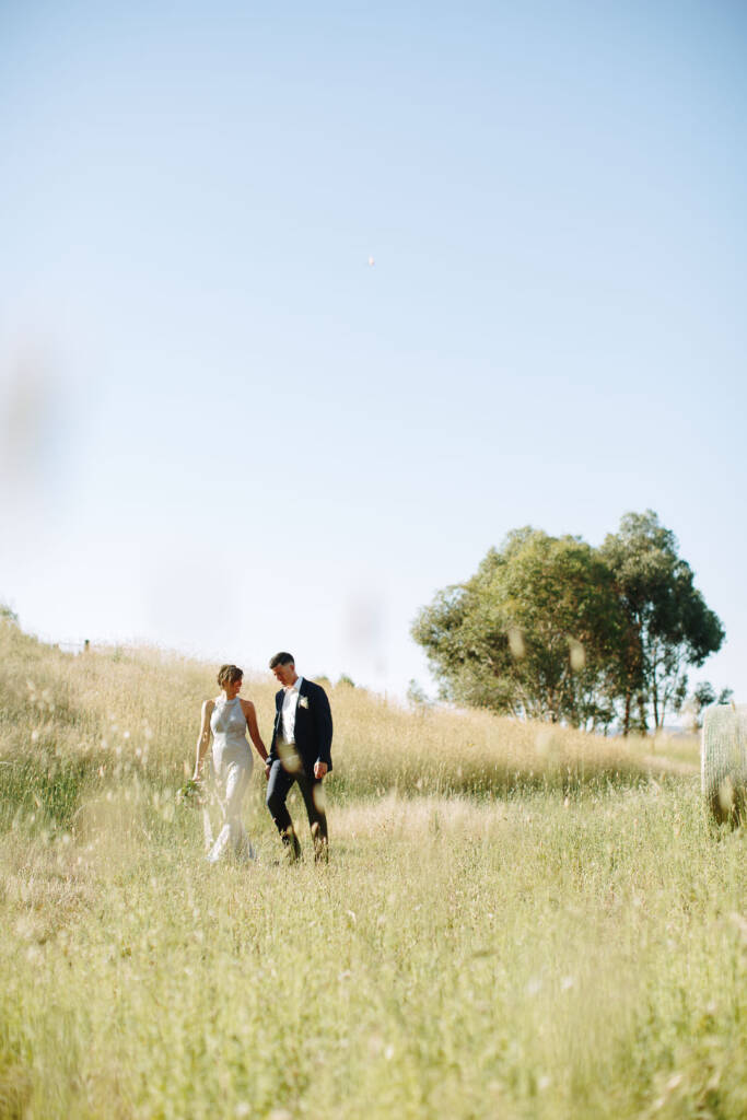 Camilla and Jason walk through tall grass beneath a tree at Flowerdale Estate.
