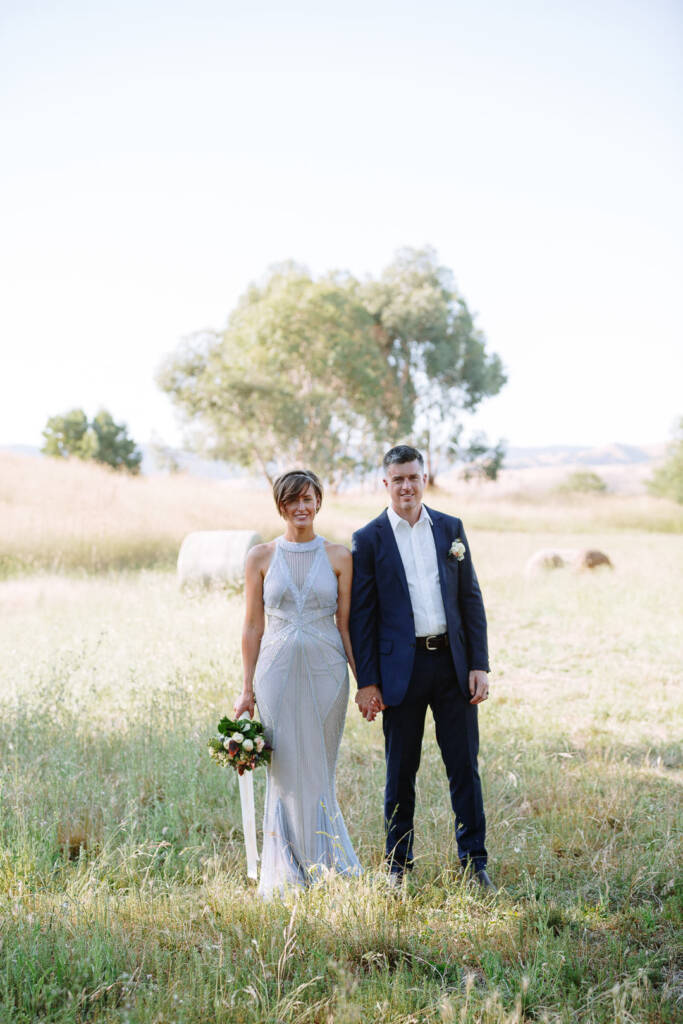 Camilla and Jason stand holding hands with hay bales behind them at Flowerdale.