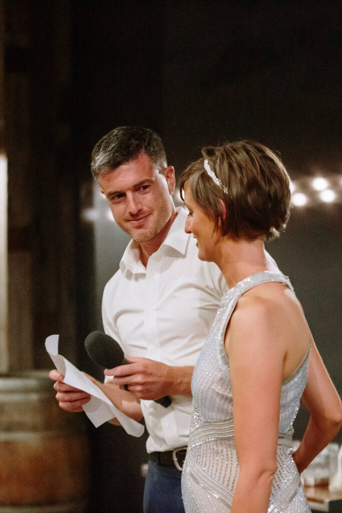 Camilla and Jason look towards each other while holding a microphone and notes.