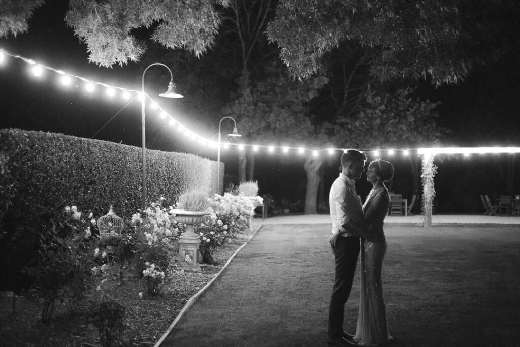 Black-and-white night portrait of Camilla and Jason beneath festoon lights in the Flowerdale garden.