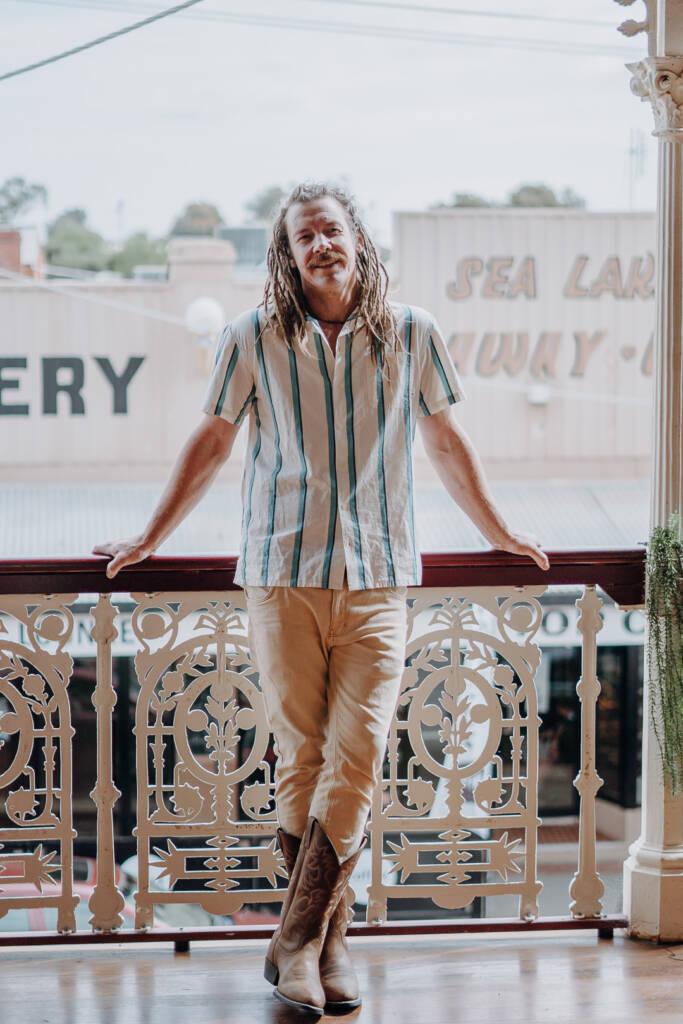 Dale leans against the Royal Hotel’s ornate balcony railing in a striped shirt and cowboy boots.