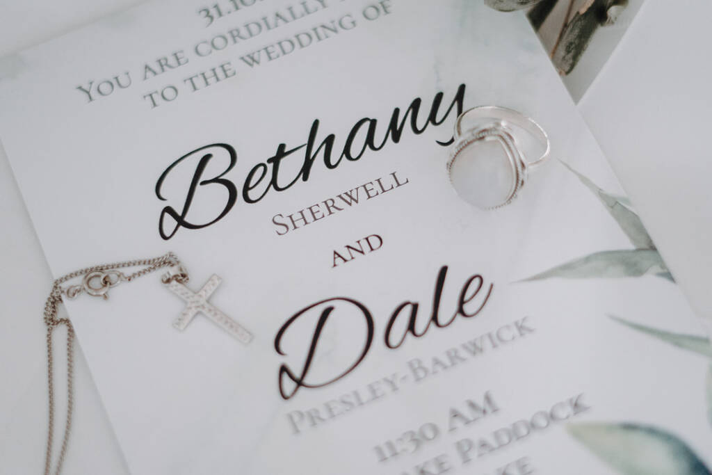 Wedding rings and a cross necklace lie across Bethany and Dale’s invitation.
