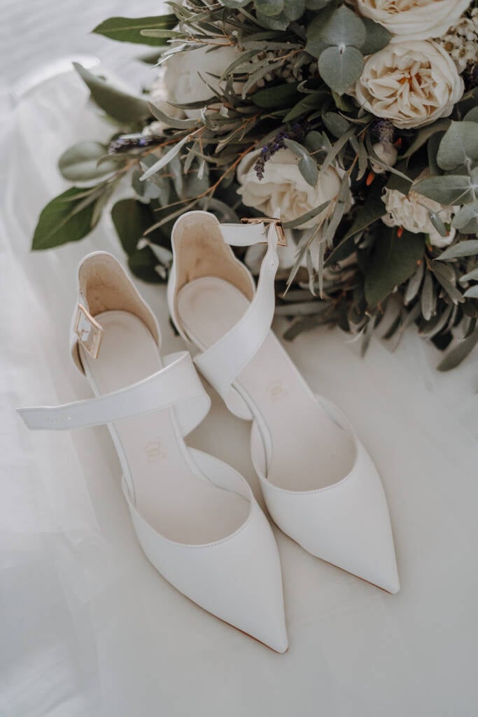White pointed slingback shoes lie beside a white-flower bouquet on pale fabric.