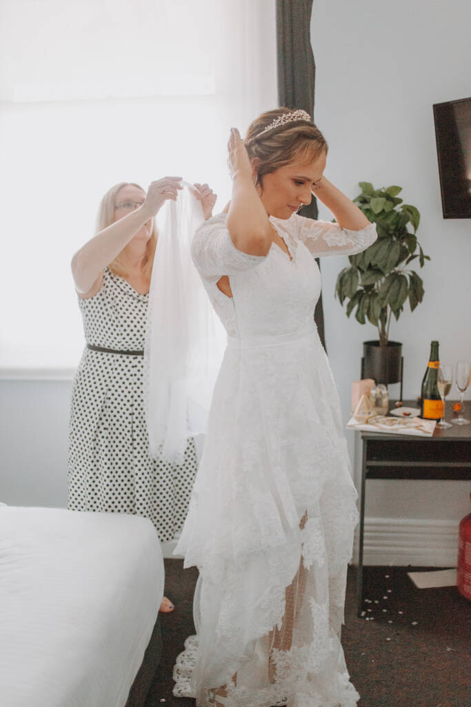 Bethany raises her hands beside her hair while a guest helps arrange her veil.