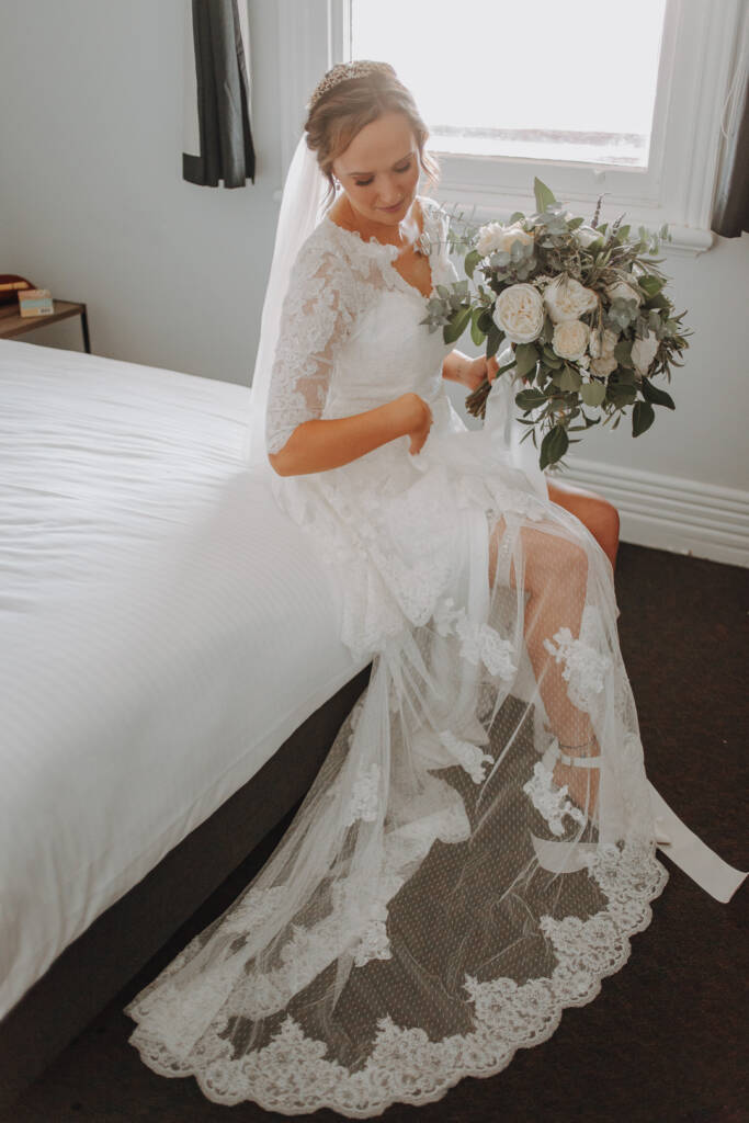Bethany sits on a bed at the Royal Hotel with her bouquet and lace train spread before her.