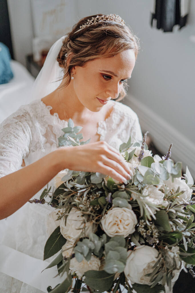 Bethany looks down over her white-flower bouquet, with her veil and jewelled hairpiece visible.