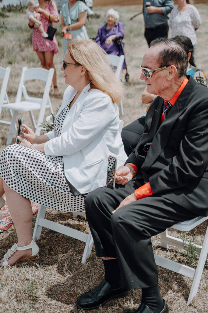 Two seated wedding guests watch the ceremony with phones in their hands.