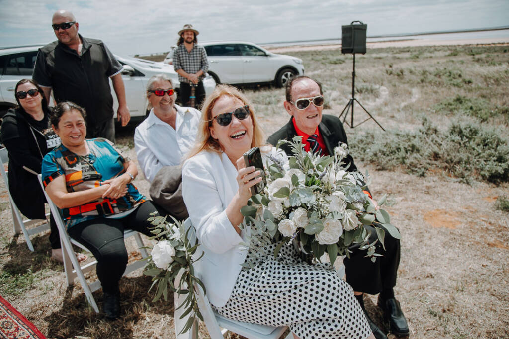 A seated guest laughs while holding a bouquet and phone during the ceremony.