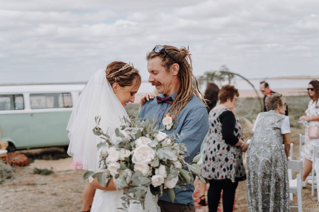 Bethany and Dale smile together while their wedding guests mingle behind them.