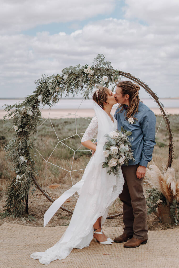 Bethany and Dale kiss beneath the boho wedding arbour at Pink Lake Reserve, Lake Tyrrell.