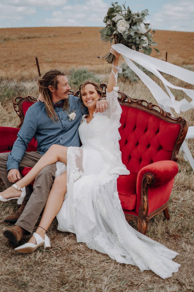 Bethany raises her bouquet while seated beside Dale on the red sofa at Lake Tyrrell.