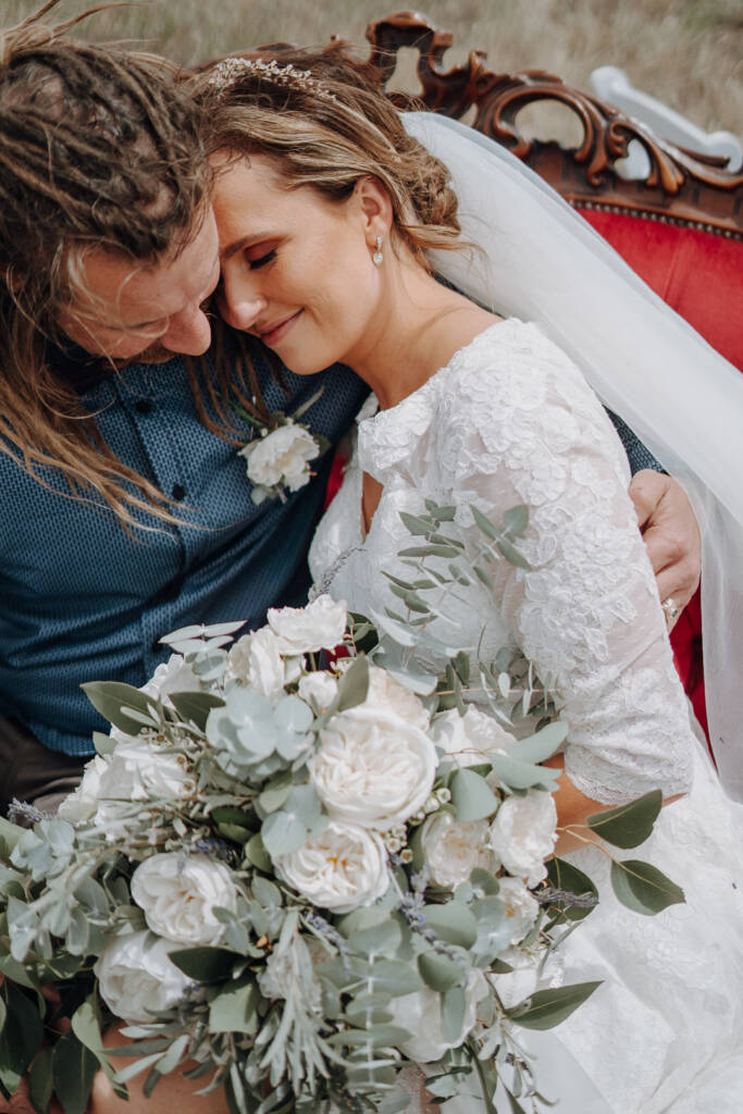 Bethany and Dale lean their heads together behind her white-flower bouquet.