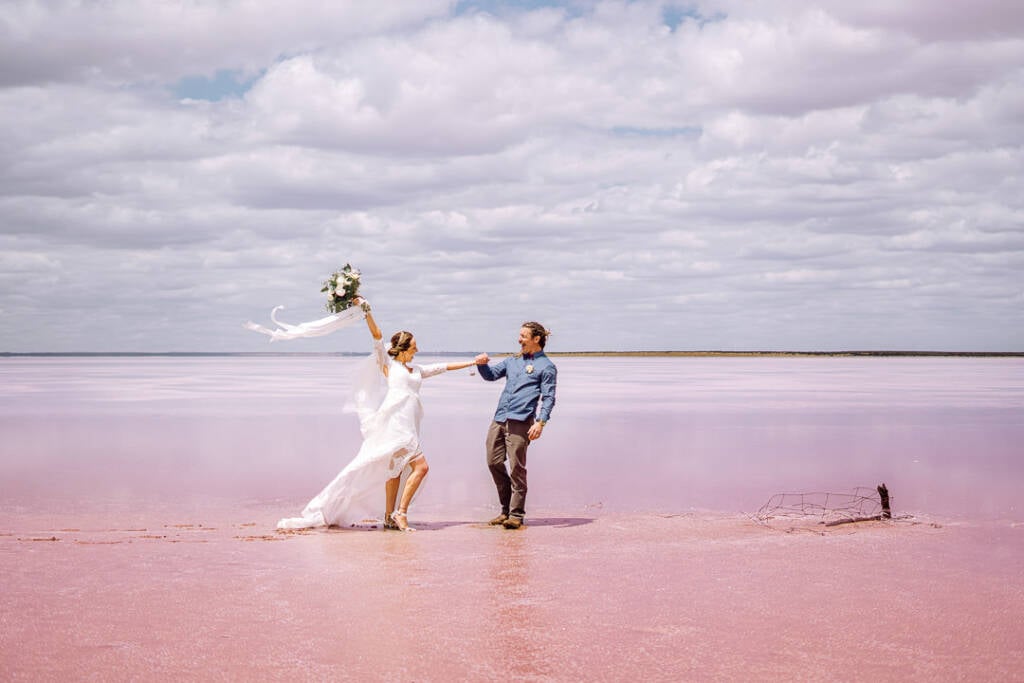 Bethany raises her bouquet while holding Dale’s hand on the pink surface of Lake Tyrrell.