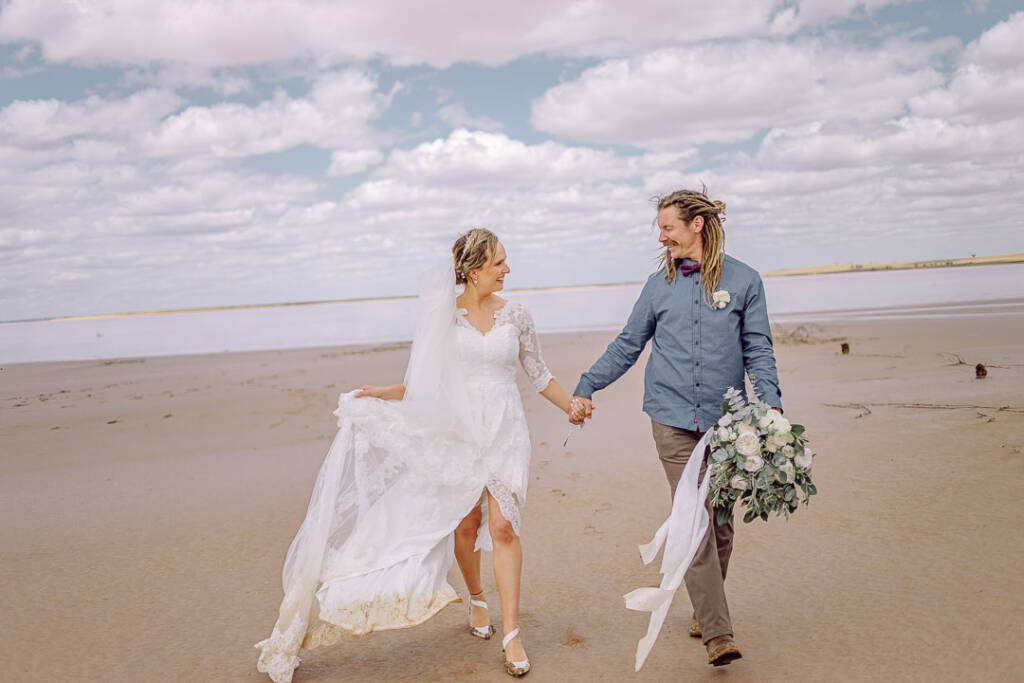 Bethany lifts her skirt while Dale carries her bouquet along Lake Tyrrell’s sandy edge.