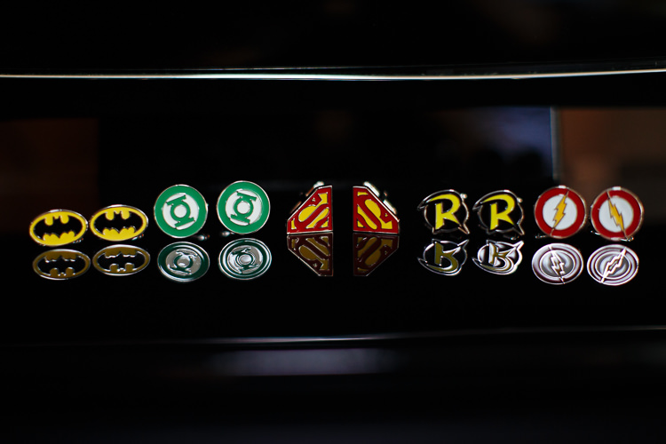 Five pairs of superhero emblem cufflinks arranged on a glossy black surface before the wedding.