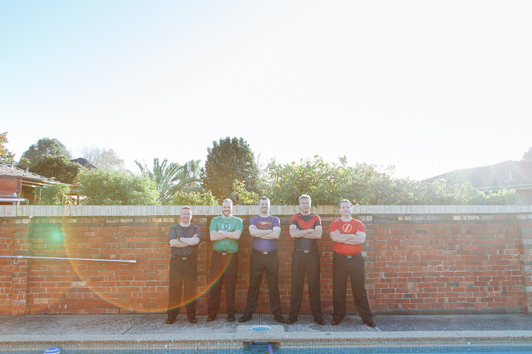 The groom and four men in superhero T shirts stand arms crossed beside a backyard pool.