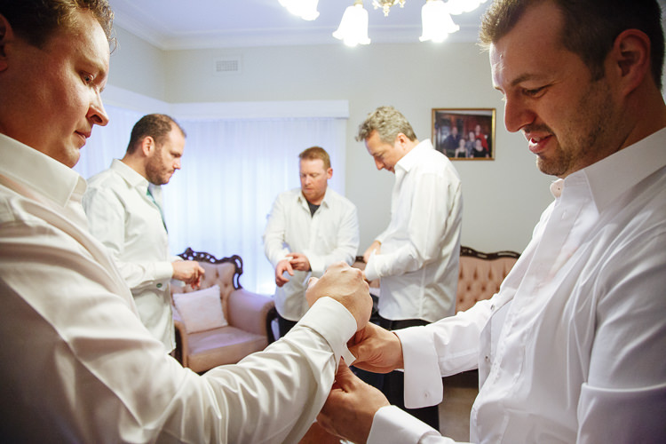A groomsman fastens the groom's cufflink as five men in white shirts get ready in the lounge.