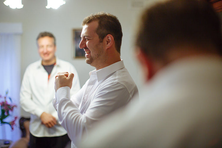 The groom laughs with a fist raised while adjusting his shirt cuff during preparation.