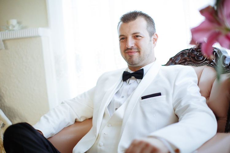The groom sits in a carved armchair in a white dinner jacket and black bow tie.