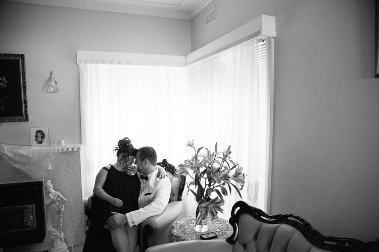The groom in the white dinner jacket sits close with a woman in black beside a bright window.