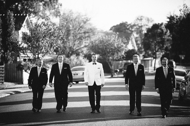 The groom in white and four groomsmen in black walk down a residential street in black and white.