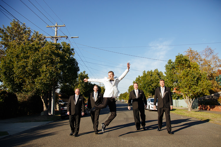 The groom leaps with one arm raised as four groomsmen in black walk beside him.