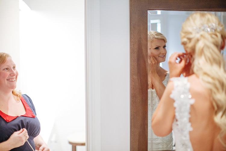 The bride fastens an earring in the timber framed mirror while the woman in navy smiles beside her.