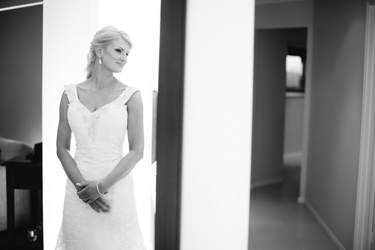 The bride stands in soft doorway light in her beaded lace gown, in black and white.